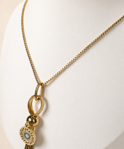 Cadena de acero quirúrgico dorado con dije de ojo turco y borla, exhibida sobre busto blanco con fondo neutro cálido.mas de cerca