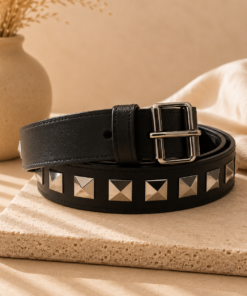 Cinto de cuero sintético con tachas piramidales metálicas y hebilla cuadrada sobre fondo beige elegante