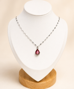 Cadena de acero quirúrgico con dije de gota verde, exhibida sobre busto blanco con fondo neutro cálido elegante.rojo