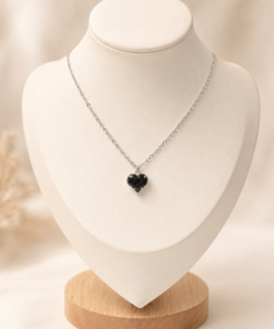 Cadena de acero quirúrgico con dije de corazón, exhibida sobre busto blanco con fondo neutro cálido elegante negro.