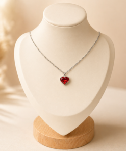 Cadena de acero quirúrgico con dije de corazón, exhibida sobre busto blanco con fondo neutro cálido elegante rojo.