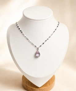 Cadena de acero quirúrgico con dije de gota verde, exhibida sobre busto blanco con fondo neutro cálido elegante rosado