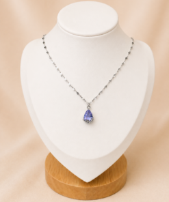 Cadena de acero quirúrgico con dije de gota verde, exhibida sobre busto blanco con fondo neutro cálido elegante.lila