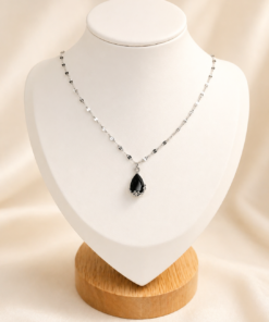 Cadena de acero quirúrgico con dije de gota verde, exhibida sobre busto blanco con fondo neutro cálido elegante.negro