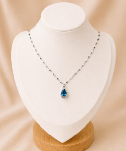 Cadena de acero quirúrgico con dije de gota verde, exhibida sobre busto blanco con fondo neutro cálido elegante.celeste