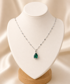 Cadena de acero quirúrgico con dije de gota verde, exhibida sobre busto blanco con fondo neutro cálido elegante verde